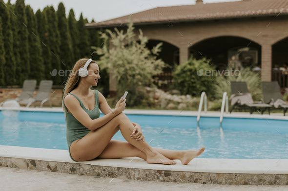 Young woman sitting by the swimming pool and listen music from mobile phone with headphones