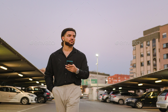 Latin man walks with a mobile phone in a parking lot at dusk