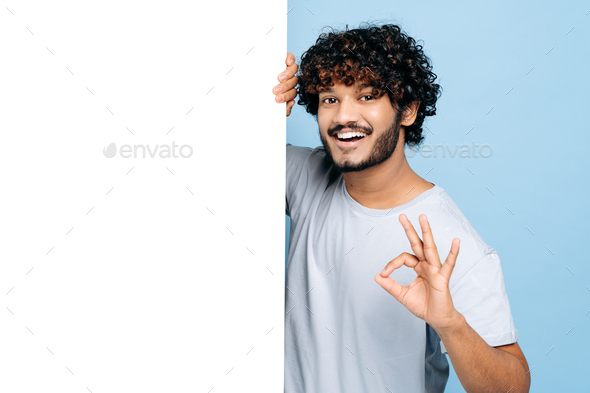 Happy Indian or Arabian guy, in casual t-shirt, peeking out from behind advertisement white board