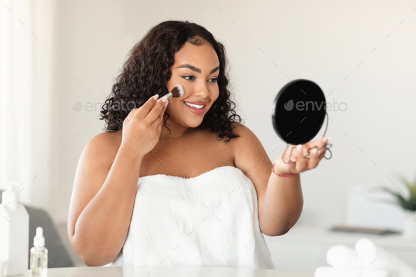 Young black oversize lady making makeup, applying facial powder with cosmetic brush, looking at