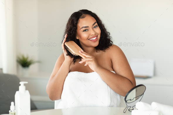 Happy black chubby woman brushing her curly hair with comb and making haircare routine at home
