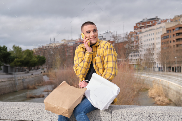 Young non binary person speaking and biting his lip on the phone with shopping bags sit on a wall