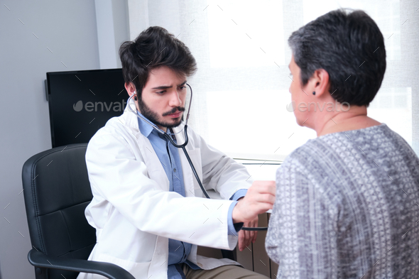 Young doctor listening to mature woman patient heart and lungs with a stethoscope.