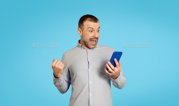 Emotional Man Holding Phone Shaking Fists Over Blue Background