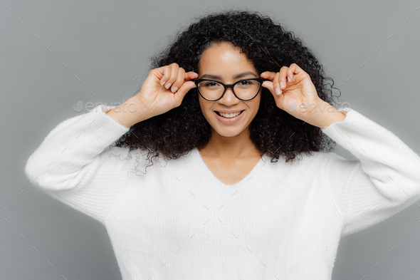 curly female with pleased facial expression, keeps hands on rim of spectacles
