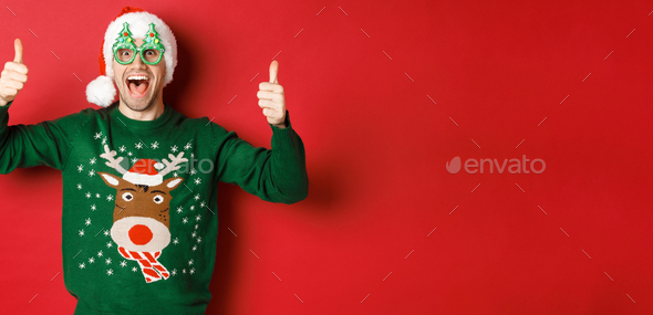Portrait of super happy young man in party glasses, santa hat and sweater, showing thumbs-up in