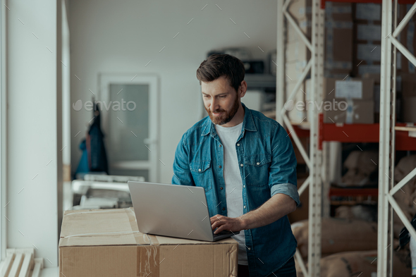 Handsome business owner works on laptop and takes order stands on own warehouse