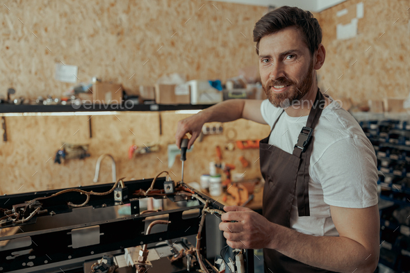 Smiling young man repairing coffee machine using screwdriver in a workshop and looking camera