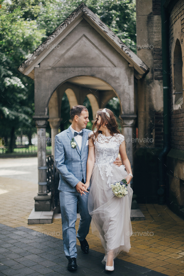 Stylish happy bride and groom walking on background of church in sunny street. Provence wedding