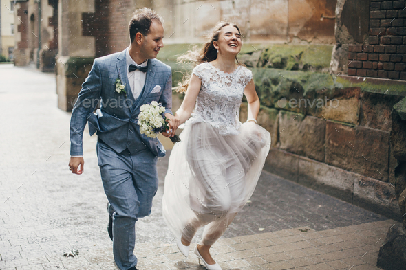 Stylish happy bride and groom running on background of old church in rainy street. Provence wedding - Stock Photo - Images