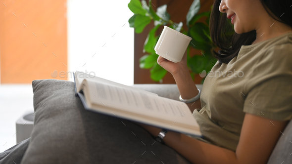 Calm asian woman drinking hot tea and reading book on comfortable couch at home.