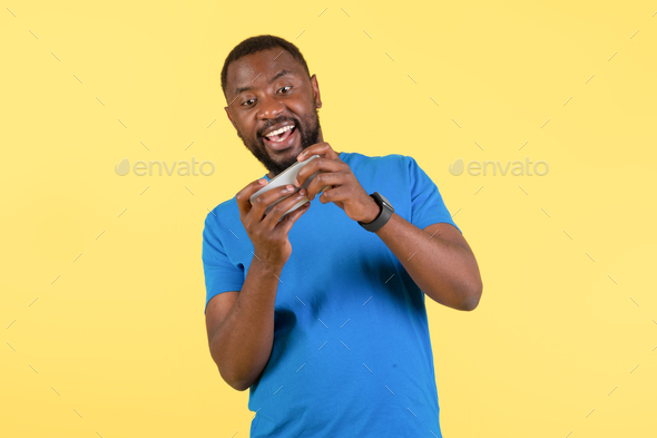 Excited Black Guy Playing Mobile Game On Cellphone, Yellow Background