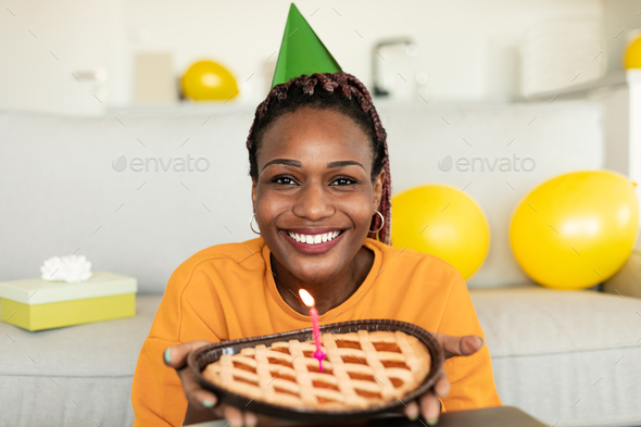 Happy birthday to you. Happy african american lady in festive hat holding pie with burning candle
