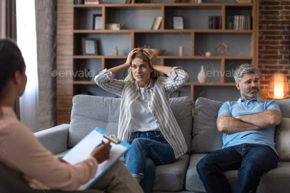 Angry middle aged caucasian wife holding her head, freaking out, husband ignores lady at meeting