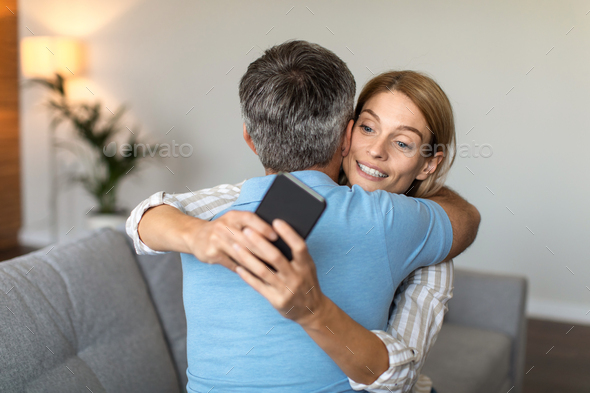 Smiling adult caucasian wife hugs husband, looks at smartphone, lady suffers from gadget addiction