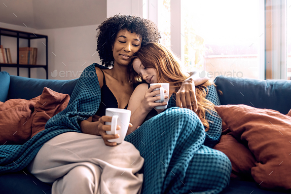 Multiethnic Lesbian Couple Hugging And Drinking Coffee On Sofa At Home