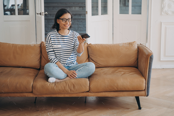 Smiling woman watching television series or movie changes TV channel, sitting on modern sofa at home