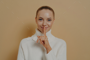 Keep it secret. Young attractive smiling lady saying be quiet with finger on lips, isolated on beige
