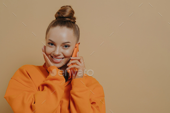 Charming young woman holding cellphone near ear, talking on phone with boyfriend, isolated on beige