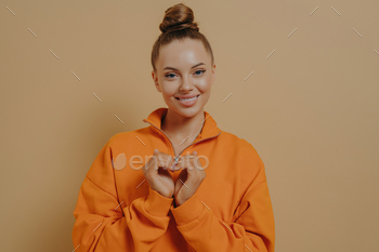 Share love. Smiling grateful young woman making heart gesture, isolated on beige studio background