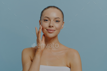 Young happy lovely woman gently touching face, having healthy pure skin, isolated on blue background