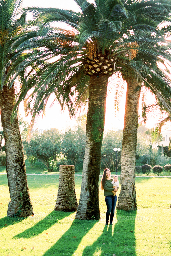 Mom with a baby in her arms stands under huge palm trees on a green lawn