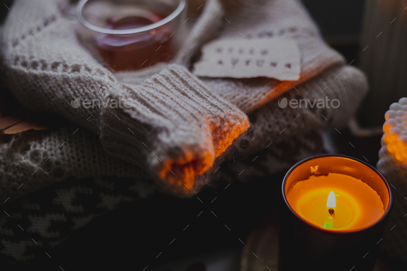Two burning candles, glass cup with tea and stack of warm cozy sweaters om bedtable. Cozy autumn