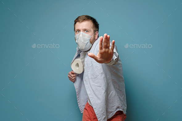 Man in a medical mask hides toilet paper under shirt and makes a stop gesture with hand.
