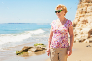 Senior woman in sunglasses stands on the beach and looks at the sea