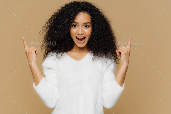 Glad young female points upwards with index fingers, shows awesome item on sale