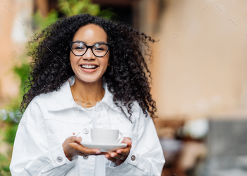 Outdoor shot of happy dark skinned lady with crisp hair, wears white jacket, drinks hot tea