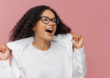 Curly overemotive woman laughs happily, keeps hands on collar of white jacket, has healthy skin