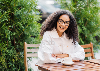 cheerful Afro woman in white clothes, drinks coffee in outdoor cafeteria, sits at wooden table