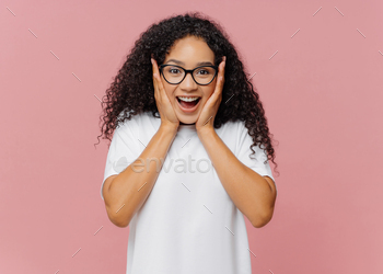 Excited African American woman touches cheeks, reacts on good news, wears transparent glasses