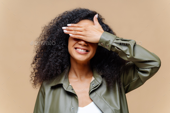 Shy cheerful young Afro woman covers eyes with palm, has toothy smile, hides face, has curly hairsty