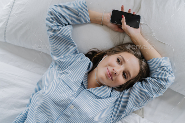 Pretty girl with pleased satisfied expression, dressed in pyjamas, rests in bed,