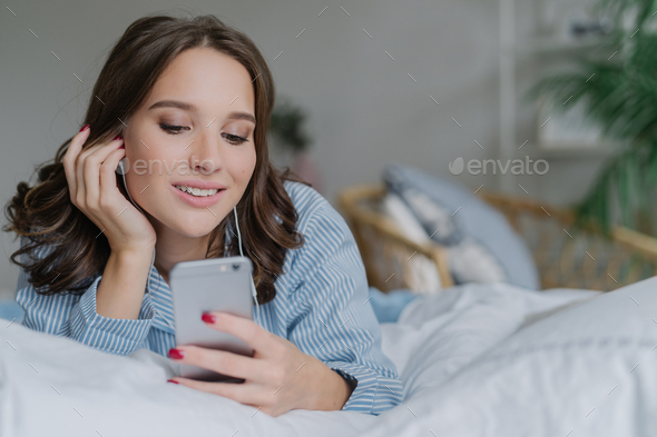 Beautiful woman listens music, makes video call, lies in comfortable bed in morning