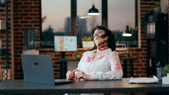 Creepy zombie sitting at desk while waving at video conference call on laptop.