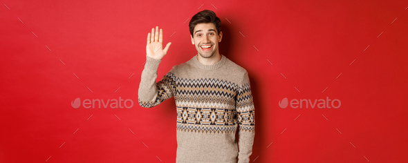 Portrait of handsome and friendly guy in christmas sweater, saying hello and waving hand, greeting