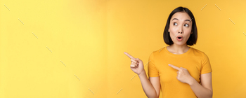 Portrait of intrigued asian woman, looking and pointing fingers left at advertisement, showing smth