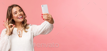 Close-up of satisfied good-looking blond girl in white dress, taking selfie, record mobile phone