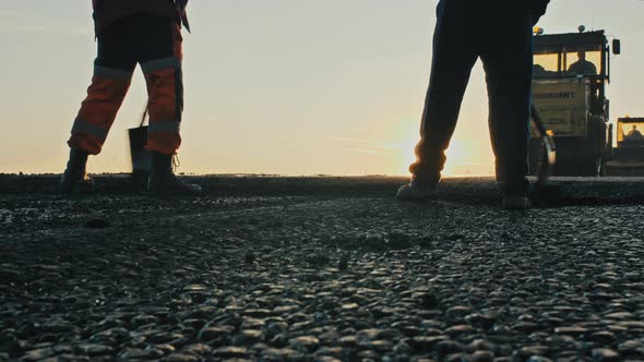 Two Road Workers in Overalls Level the Fresh Asphalt Mix Against the Background of Road Rollers