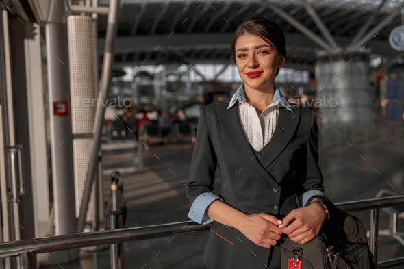 Smiling beautiful air hostess holding bag and posing at camera