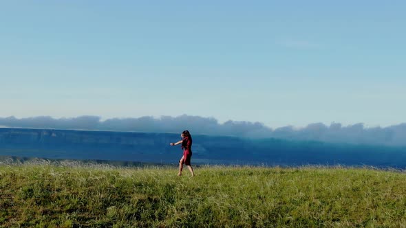Aerial View of a Young Barefoot Woman in a Red Dress Who Dances in the Grass Against the Background alt