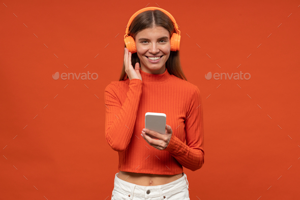 Woman music fan in wireless headphones holding phone listening to classic music on orange background