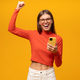 Lucky excited woman holding smartphone making winner gesture shouting yes on yellow background - PhotoDune Item for Sale