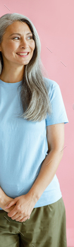 Happy silver haired middle aged Asian woman in t-shirt stands on pink background
