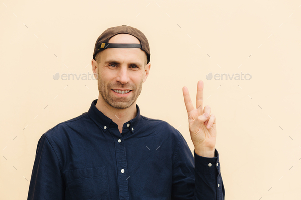 Portrait of handsome delighted unshaven male shows peace sign, keeps two fingers raised