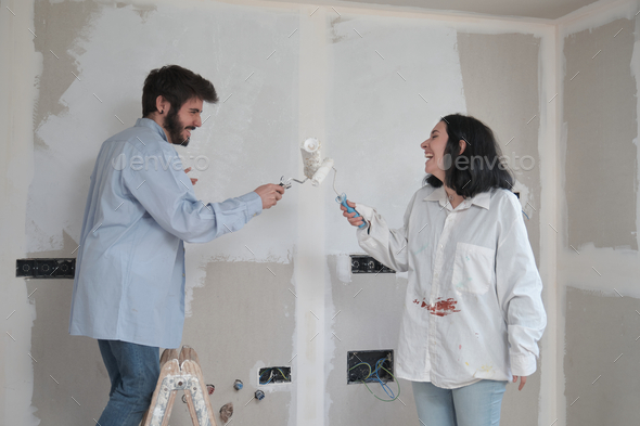 Young couple play sword fight with paint rollers and laugh while painting walls.