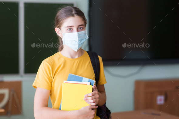 portrait of student in medical mask with backpack, notebooks against background of blackboard.